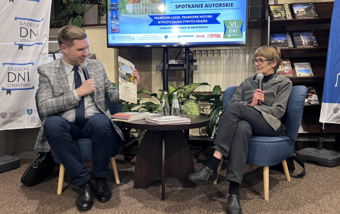 Dwie osoby siedzą na niskich niebieskich fotelach. Jedną z tych osób jest uśmiechnięty mężczyzna w szarej marynarce w kratę. Trzyma w jeden ręce mikrofon, a w drugiej książkę. Obok niego siedzi kobieta w krótkich jasnych włosach w szarym stroju i czarnych okularach. Trzyma mikrofon w ręce. Pomiędzy nimi jest niski drewniany stolik. Na stolik leży stosik książek i szklane butelki. Za nimi rośliny i duży ekran z napisem: Marzanna Raińska - spotkanie autorskie.