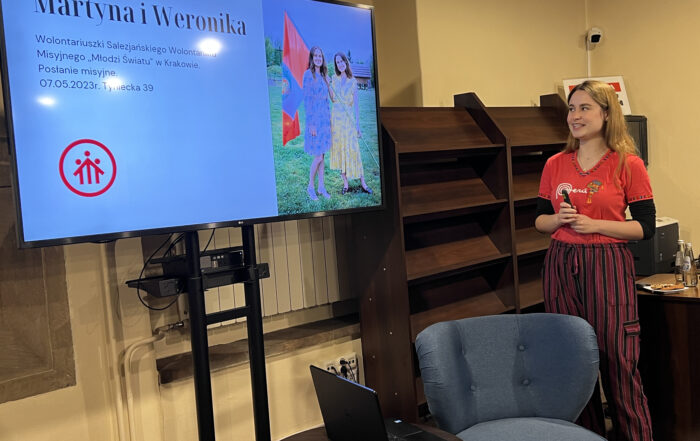 Młoda kobieta w długich prostych włosach. Ma na sobie czerwoną koszulkę i ciemne spodnie w fioletowe paski. Uśmiecha się. Przed nią znajduje się niski ciemny stolik. Na nim stoi laptop. Obok jest niskie niebieskie krzesło. Nad stolikiem znajduje się duży ekran ze zdjęciem. Na zdjęciu dwie uśmiechnięte dziewczyny, które trzymają w ręce dużą flagę. Bok nich napis: Martyna i Weronika.