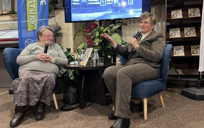 Na zdjęciu widać dwie kobiety siedzące na krzesłach. Kobieta po lewej stronie ma na sobie szary sweter i wzorzystą spódnicę, trzyma mikrofon i patrzy w lewo. Kobieta po prawej stronie ubrana jest w brązowy garnitur, również trzyma mikrofon i gestykuluje ręką, jakby mówiła. W tle, za kobietami, znajduje się ekran telewizora, na którym wyświetlane jest czarno-białe zdjęcie starego drewnianego domu. Przed ekranem stoi stolik z kwiatami w wazonie i butelkami wody. Po prawej stronie widoczny jest regał z książkami. W lewym górnym rogu wisi baner.