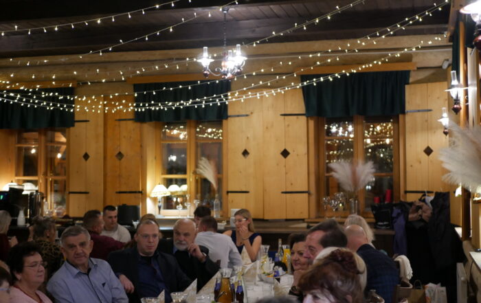 Sala bankietowa z kilkudziesięcioma osobami siedzącymi przy stołach. Pod drewnianym sufitem rozwieszone lampki. W oknach stoją zapalone lampki z jasnymi kloszami oraz flakony z białymi długimi piórami.