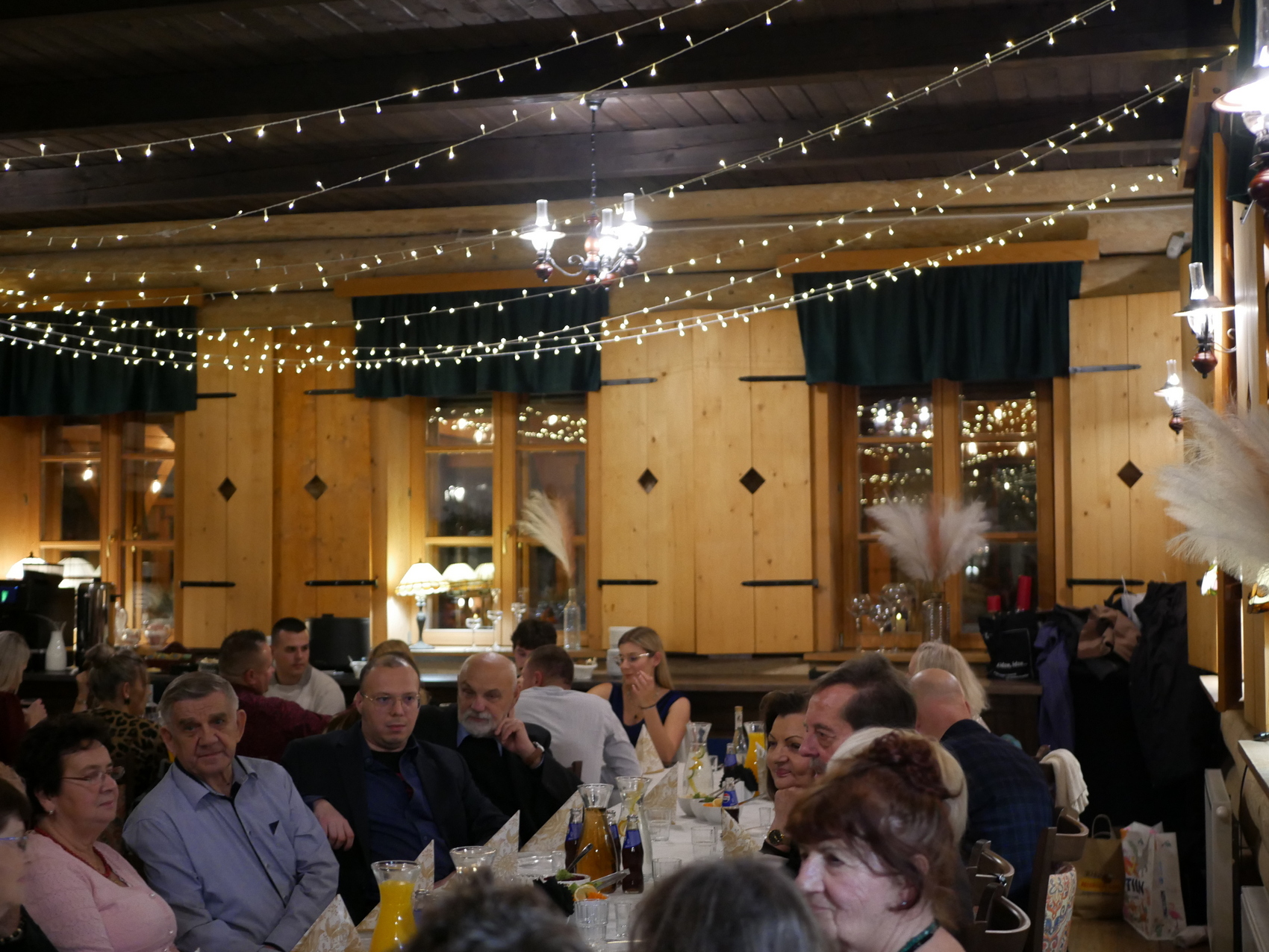 Sala bankietowa z kilkudziesięcioma osobami siedzącymi przy stołach. Pod drewnianym sufitem rozwieszone lampki. W oknach stoją zapalone lampki z jasnymi kloszami oraz flakony z białymi długimi piórami.