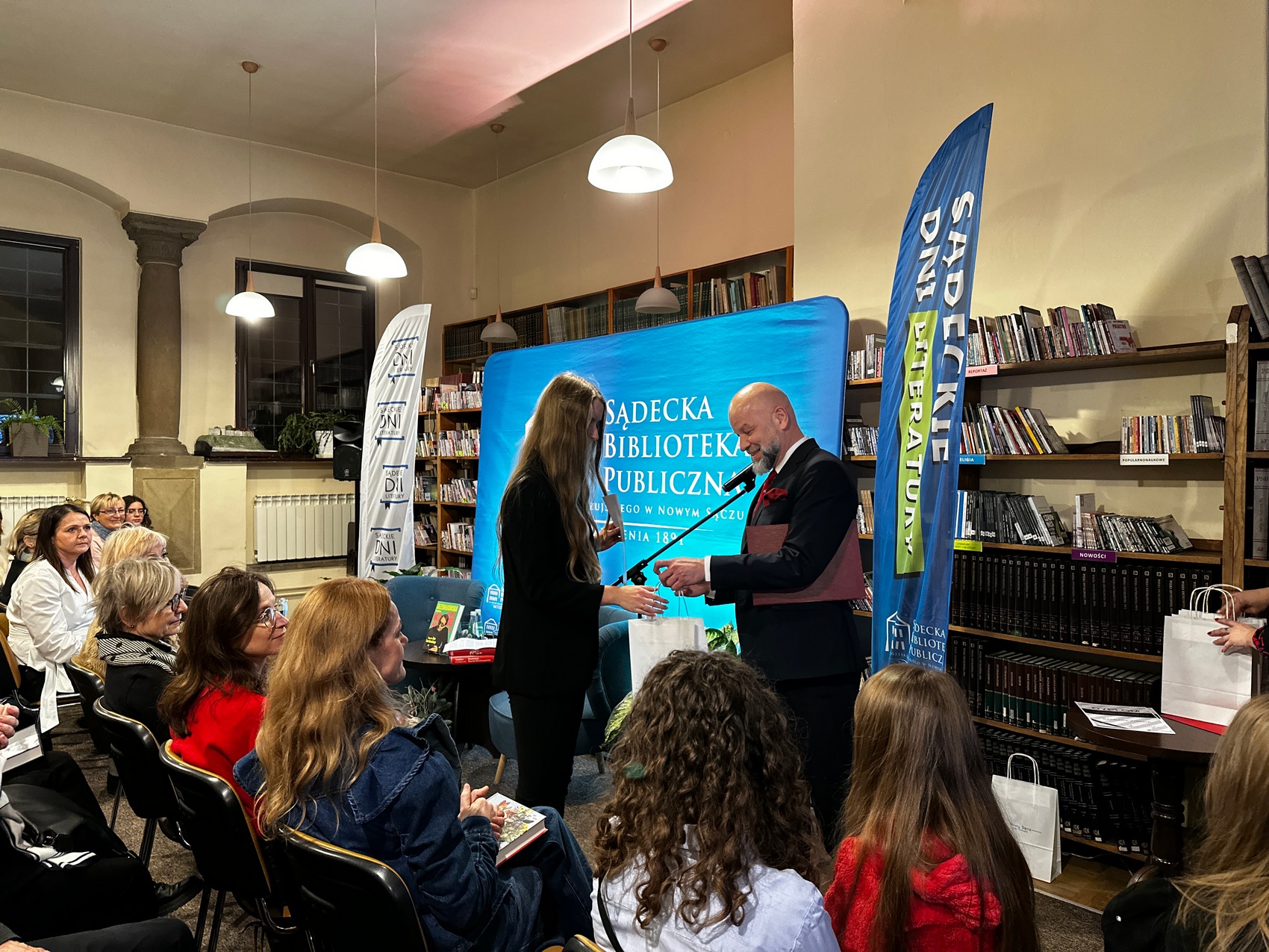 Dyktando_1 Mężczyzna w granatowym garniturze, białej koszuli i bordowym krawacie wręcza białą torbę prezentową ubranej w czarną garsonkę kobiecie z długimi blond włosami. Za nimi błękitna ścianka reklamowa z białym logotypem Sądeckiej Biblioteki Publicznej, dwie flagi reklamowe i regał z książkami. Przed nimi siedząca na krzesłach publika.