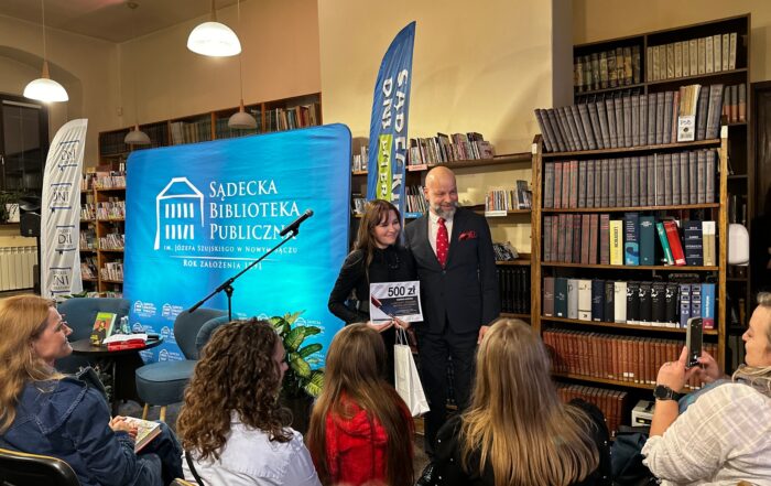 Mężczyzna w granatowym garniturze, białej koszuli i bordowym krawacie stoi obok ubranej w czarną sukienkę kobiety. Kobieta trzyma w rękach dyplom i białą torbę prezentową.Za nimi błękitna ścianka reklamowa z białym logotypem Sądeckiej Biblioteki Publicznej, dwie flagi reklamowe i regał z książkami. Przed nimi siedząca na krzesłach publika.