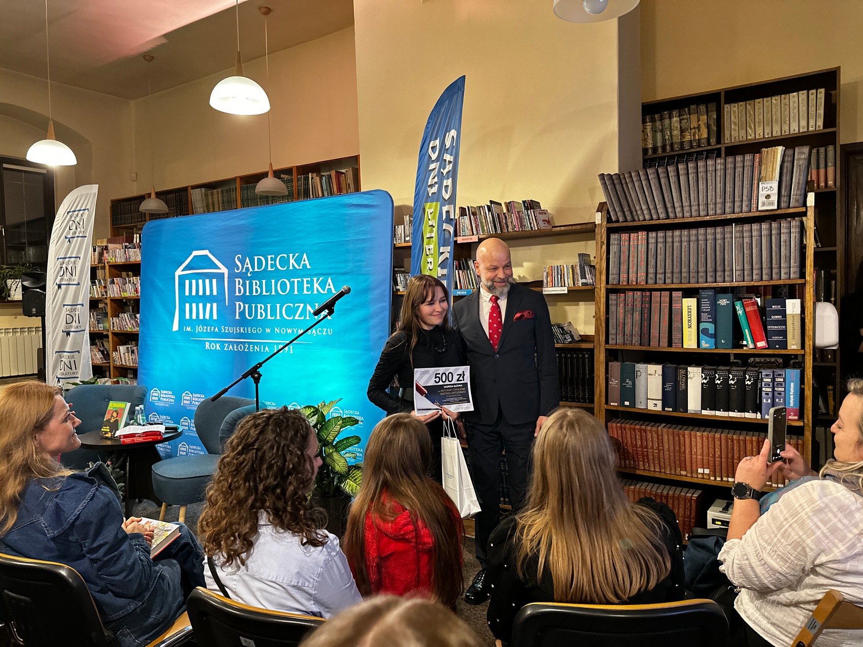 Mężczyzna w granatowym garniturze, białej koszuli i bordowym krawacie stoi obok ubranej w czarną sukienkę kobiety. Kobieta trzyma w rękach dyplom i białą torbę prezentową.Za nimi błękitna ścianka reklamowa z białym logotypem Sądeckiej Biblioteki Publicznej, dwie flagi reklamowe i regał z książkami. Przed nimi siedząca na krzesłach publika.