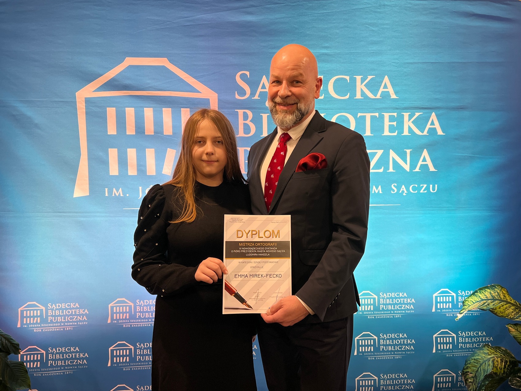 Dyktando_3 Mężczyzna w granatowym garniturze, białej koszuli i bordowym krawacie stoi obok ubranej w czarną sukienkę kobiety. Trzymają w rękach dyplom i uśmiechają się. Za nimi błękitna ścianka reklamowa z białym logotypem Sądeckiej Biblioteki Publicznej.