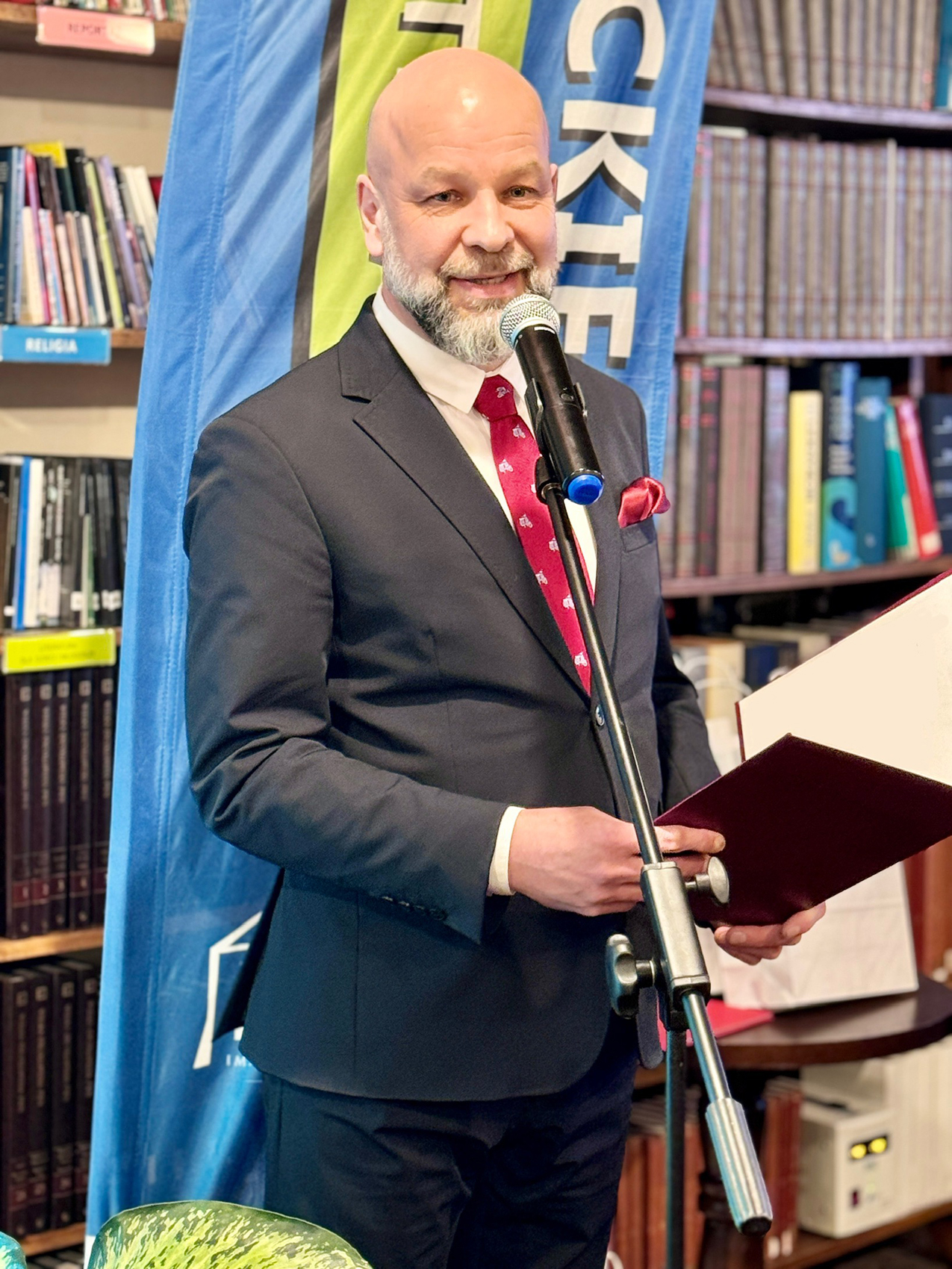 Dyktando_4 Mężczyzna w granatowym garniturze, białej koszuli i bordowym krawacie mówi do ustawionego na statywie mikrofonu. W tle kolorowa flaga reklamowa oraz regał z książkami.