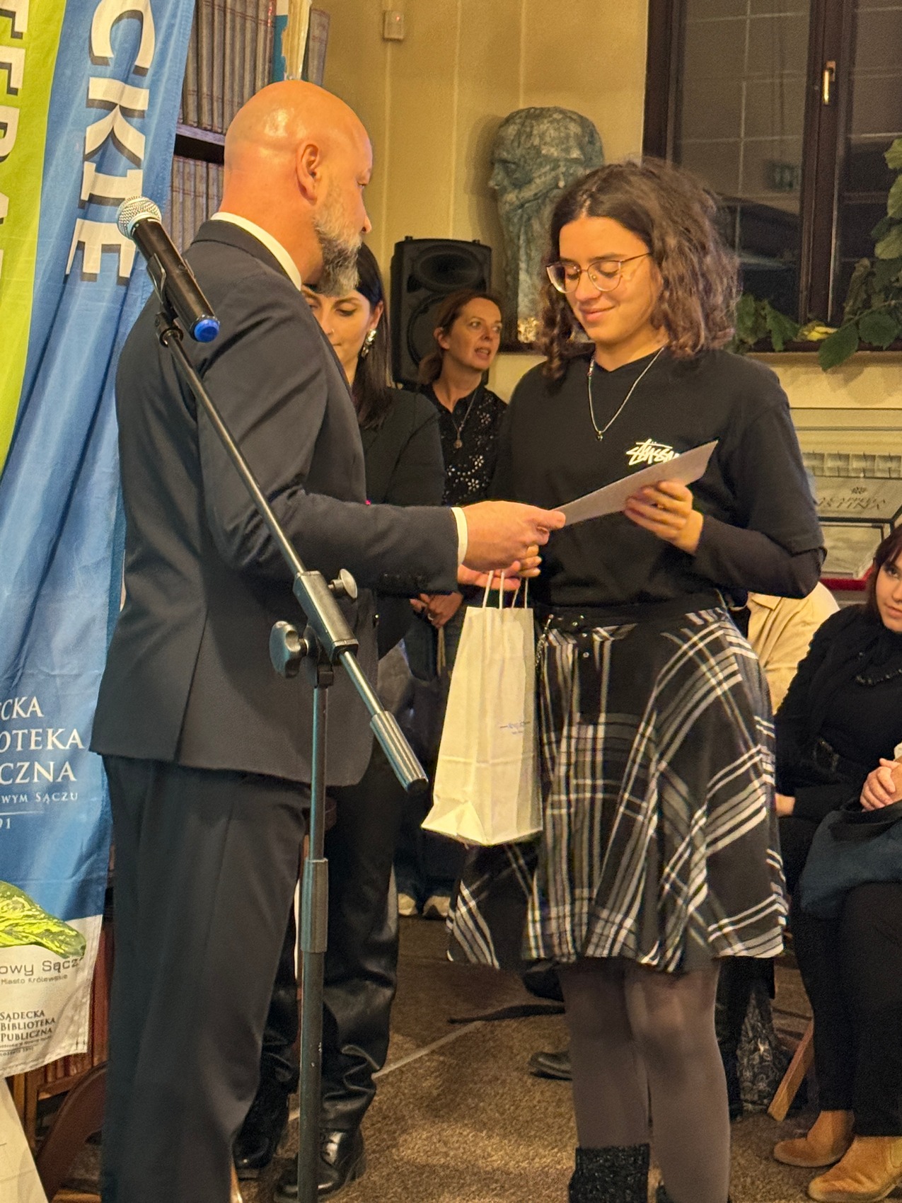 Dyktando_5 Mężczyzna w granatowym garniturze wręcza ubranej w kraciastą czarno-białą spódnicę kobiecie dyplom i torbę prezentową. Obydwoje się uśmiechają. W tle kolorowa flaga reklamowa oraz regał z książkami.