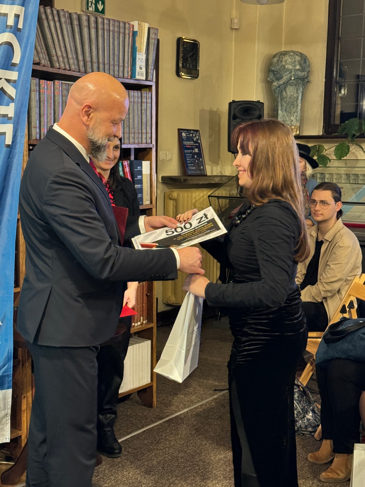 Dyktando_6 Mężczyzna w granatowym garniturze wręcza ubranej w czarną sukienkę kobiecie dyplom z napisem pięćset złotych. Obydwoje się uśmiechają. W tle regał z książkami.