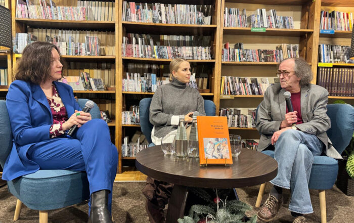 W pomieszczeniu na niebieskich fotelach siedzą trzy osoby: dwie kobiety i mężczyzna. Kobieta od lewej ubrana jest w niebieski garnitur. W rękach opartych na kolanach trzyma mikrofon. Obok niej, pośrodku, siedzi druga kobieta ubrana w szary golf. Po prawej stronie siedzi starszy mężczyzna ubrany w szarą marynarkę i jeansy. W ręce trzyma mikrofon blisko ust, mówi do niego. Mężczyzna patrzy na kobiety, a kobiety patrzą na mężczyznę. Przed nimi stoi mały okrągły brązowy stolik. Na stoliku znajdują się: dwie szklanki, dwie butelki z wodą i książka z pomarańczową okładką. Pod stolikiem stoi świąteczny stroik. W tle za siedzącymi stoją wysokie regały z audiobookami.