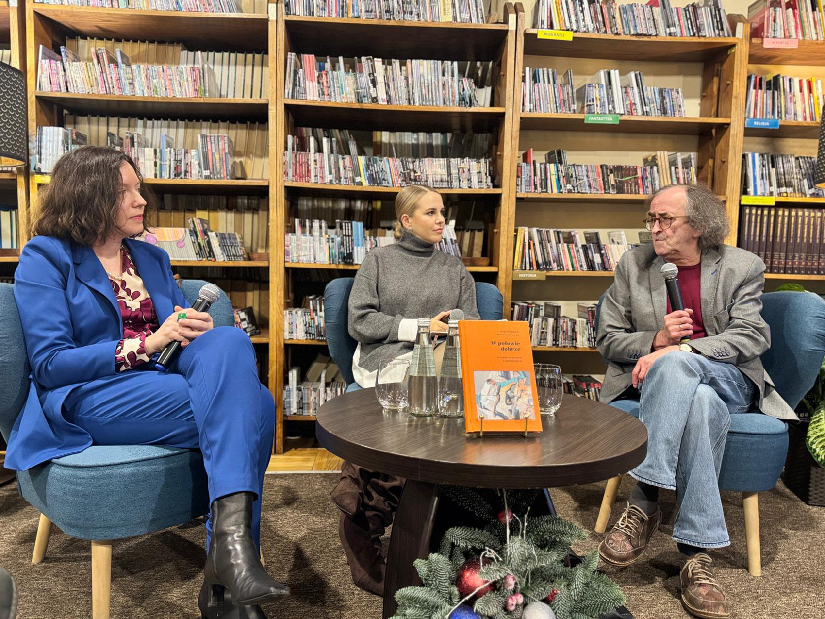W pomieszczeniu na niebieskich fotelach siedzą trzy osoby: dwie kobiety i mężczyzna. Kobieta od lewej ubrana jest w niebieski garnitur. W rękach opartych na kolanach trzyma mikrofon. Obok niej, pośrodku, siedzi druga kobieta ubrana w szary golf. Po prawej stronie siedzi starszy mężczyzna ubrany w szarą marynarkę i jeansy. W ręce trzyma mikrofon blisko ust, mówi do niego. Mężczyzna patrzy na kobiety, a kobiety patrzą na mężczyznę. Przed nimi stoi mały okrągły brązowy stolik. Na stoliku znajdują się: dwie szklanki, dwie butelki z wodą i książka z pomarańczową okładką. Pod stolikiem stoi świąteczny stroik. W tle za siedzącymi stoją wysokie regały z audiobookami.