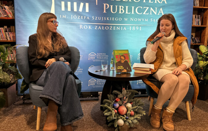 Dwie kobiety siedzą na niskich niebieskich fotelach. Kobieta lewej ma długie brązowe włosy i okulary. Ubrana jest w ciemne jeansy i czarną marynarkę. Trzyma w ręce mikron. Druga z kobiet ma na sobie sweterkową sukienkę, żółtą kamizelę. Trzyma w ręce mikrofon przy ustach. Pomiędzy nimi stoi ciemny okrągły stolik. Na nim książki i szklanka wody. pod stolikiem mały stroik bożonarodzeniowy z kolorowymi bombkami. Za nimi niebieska ścianka.