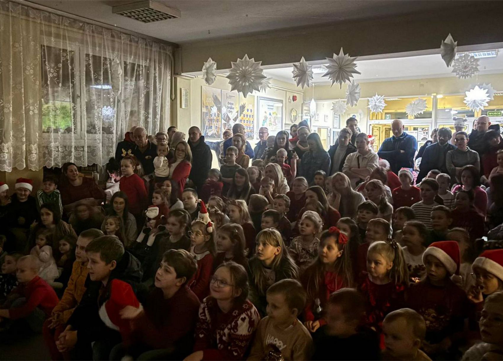 Mikołajki BO_2 Pełna sala dzieci i osób dorosłych. Część z nich ma na głowach czapki mikołajkowe. Sala wystrojona świątecznie - pod sufitem wiszą duże białe gwiazdy.