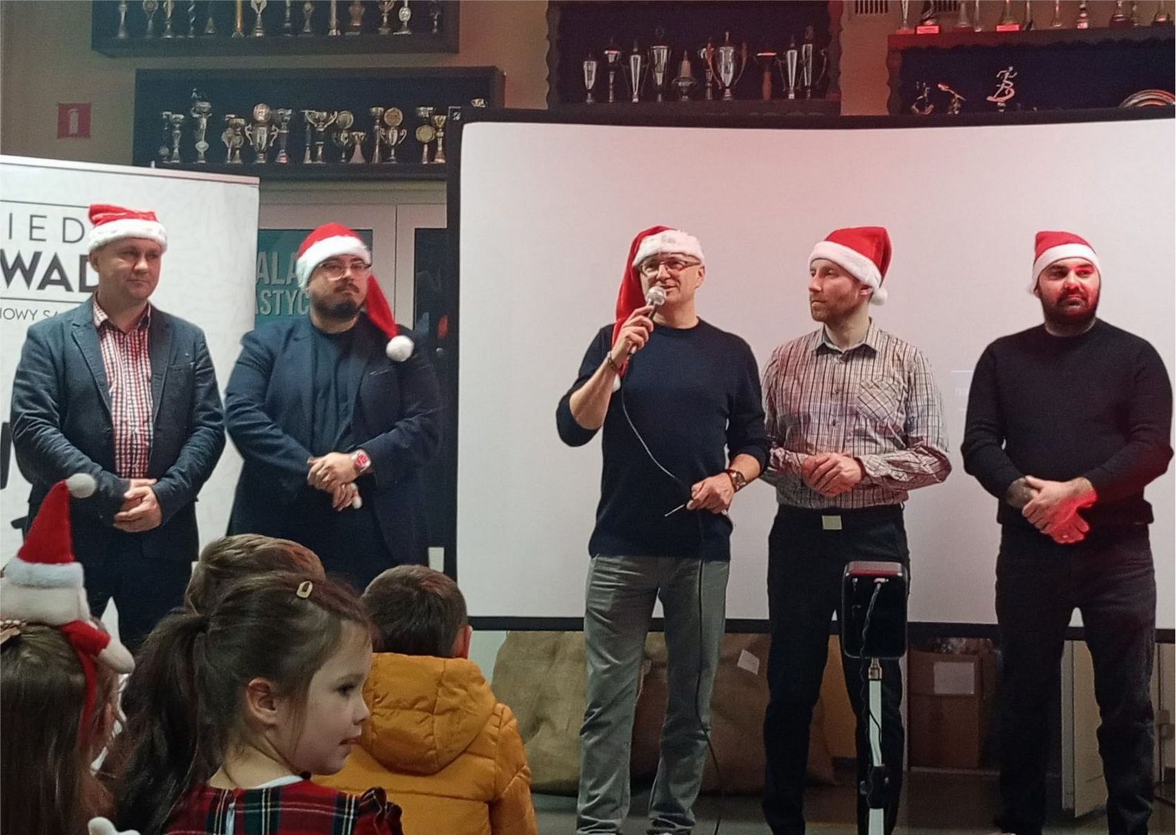 Mikołajki_BO_1 Pięciu mężczyzn w mikołajkowych czapkach stoi przed siedzącymi dziećmi. Jeden z nich ma w ręce mikrofon. Wszyscy mają ubrane mikołajkowe czapki.