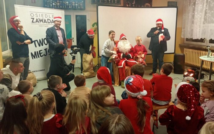 Na froncie siedzi Mikołaj, przy nim stoi dziewczynka i chłopiec. Obok nich czterech mężczyzn w mikołajkowych czapkach. Wszyscy stoją zwróceni w stronę pełnej sali dzieci.