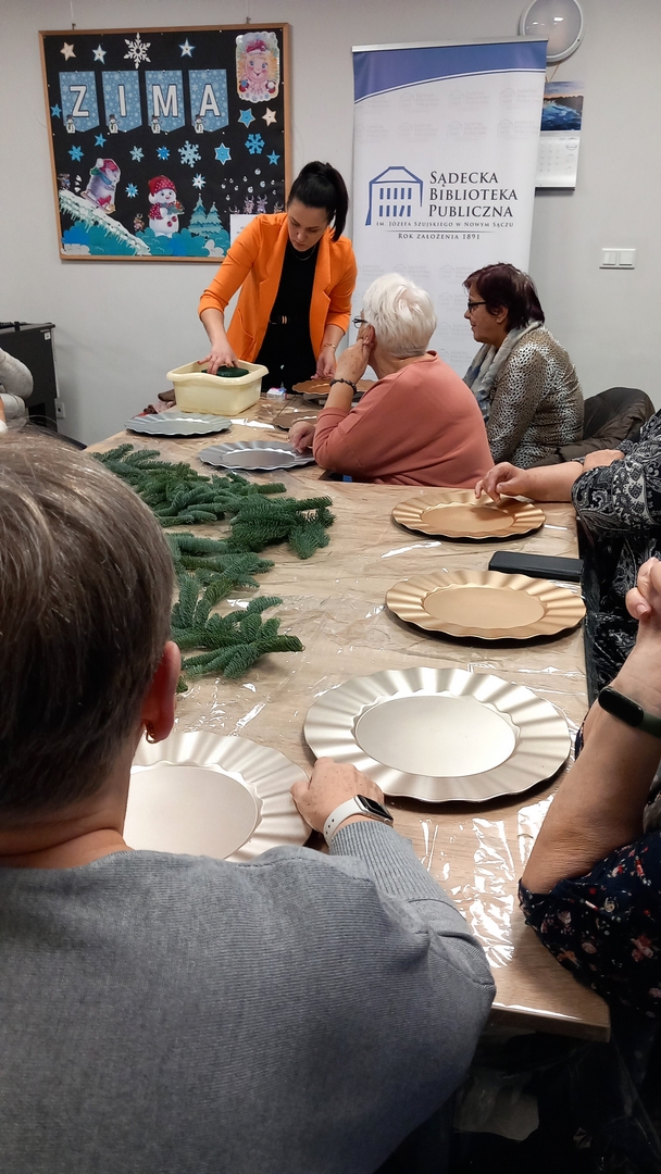 florystyczne02 Wokół stołu siedzi kilka osób, słuchają, patrzą na stojącą pośrodku kobietę w pomarańczowej marynarce. Kobieta pochyla się nad miską z wodą i gąbką florystyczną, tłumaczy coś. Na stole leżą złote i srebrne tacki oraz zielone gałązki jedliny.