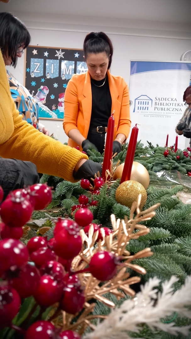 florystyczne10 Wnętrze, na stole stroiki z żywej jedliny. Wokół kilka osób, widać ich dłonie. Pośrodku, przy stole stoi kobieta w pomarańczowej marynarce. Na pierwszym planie duży stroik z zielonych gałązek, z dwoma czerwonymi świeczkami, dużymi złotymi bombkami i złotymi gałązkami.