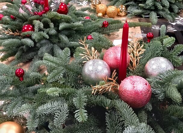 Wnętrze, na połączonych pośrodku stołach stroiki świąteczne. W tle widać fragmenty dwóch okien oraz dłonie przypinające ozdoby. Stroiki z żywej jedliny, z białymi i czerwonymi świeczkami, srebrnymi, czerwonymi, złotymi i różowymi bombkami. Pomiędzy stroikami leżą gałązki, szyszki i bombki.