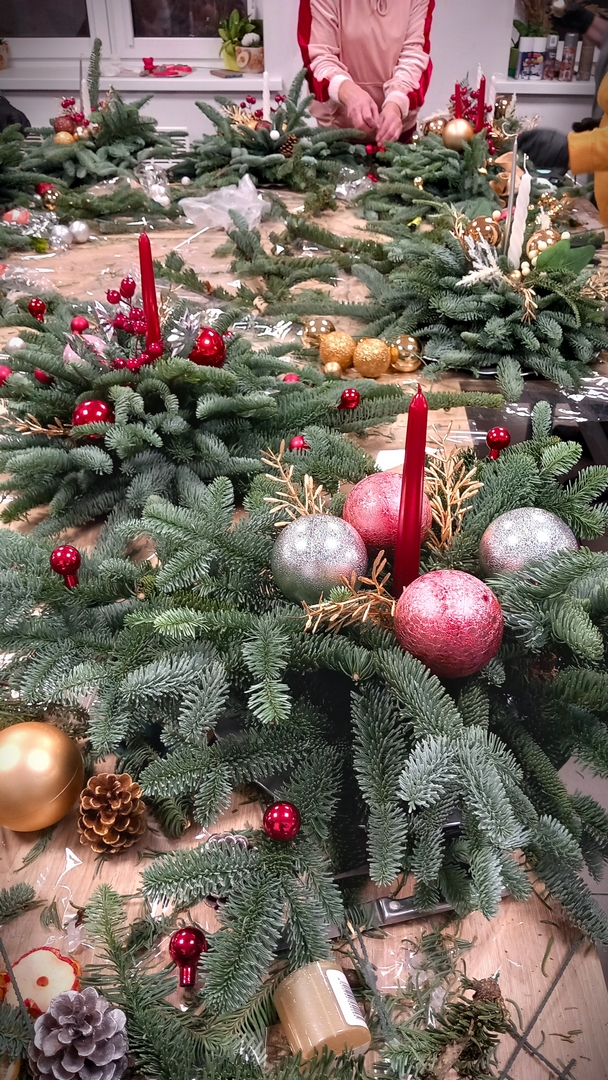 florystyczne15 Wnętrze, na połączonych pośrodku stołach stroiki świąteczne. W tle widać fragmenty dwóch okien oraz dłonie przypinające ozdoby. Stroiki z żywej jedliny, z białymi i czerwonymi świeczkami, srebrnymi, czerwonymi, złotymi i różowymi bombkami. Pomiędzy stroikami leżą gałązki, szyszki i bombki.