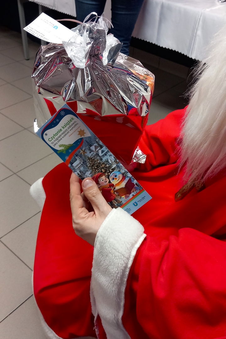 mikolajki03 Zbliżenie na dłonie Świętego Mikołaja. W jednej trzyma prezent w srebrnym worku, a w drugiej zakładkę ze świątecznym obrazkiem i napisem. Na obrazku wiele domków z dachami pokrytymi śniegiem, dużą choinką. Na pierwszym planie bajkowy chłopiec w czerwonej kurtce i czapce oraz bałwanek trzymający otwartą książkę. Cytat na górze zakładki: Czytanie książek to najpiękniejsza zabawa jaką sobie ludzkość wymyśliła - Wisława Szymborska.