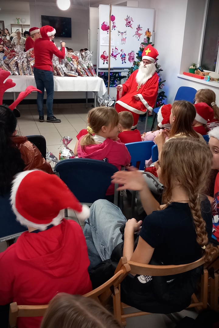 mikolajki06 Wnętrze świątecznie udekorowane. W głębi pomieszczenia, na stole przykrytym białym obrusem położonych jest wiele paczek w srebrnych woreczkach. Obok stołu, przy choince, siedzi Święty Mikołaj oraz stoi kobieta w czerwonym swetrze i czerwonej czapce z pomponem. Przed nimi na krzesłach siedzą dzieci. Wiele z nich ma na głowach czerwone czapki z białymi pomponami.