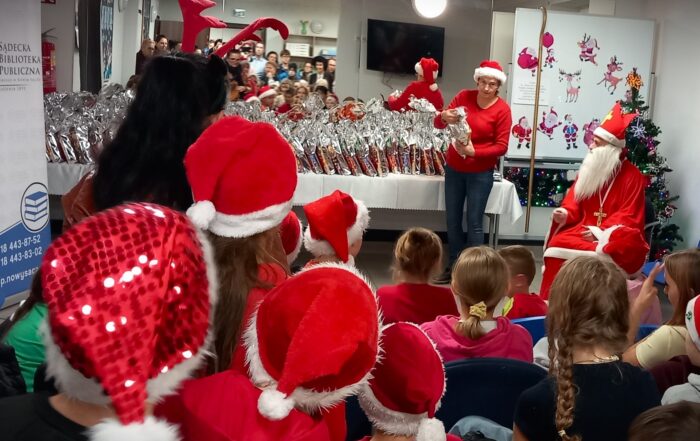 Wnętrze świątecznie udekorowane. W głębi pomieszczenia, na stole przykrytym białym obrusem położonych jest wiele paczek w srebrnych woreczkach. Obok stołu, przy choince, siedzi Święty Mikołaj oraz stoi kobieta w czerwonym swetrze i czerwonej czapce z pomponem. Przed nimi na krzesłach siedzą dzieci. Wiele z nich ma na głowach czerwone czapki z białymi pomponami. Po lewej stronie stoi rollup z logotypem Sądeckiej Biblioteki Publicznej.
