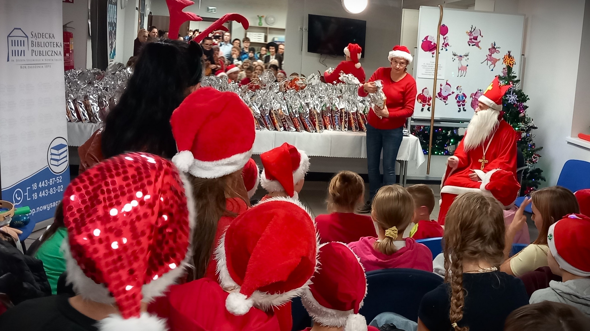mikolajki08 Wnętrze świątecznie udekorowane. W głębi pomieszczenia, na stole przykrytym białym obrusem położonych jest wiele paczek w srebrnych woreczkach. Obok stołu, przy choince, siedzi Święty Mikołaj oraz stoi kobieta w czerwonym swetrze i czerwonej czapce z pomponem. Przed nimi na krzesłach siedzą dzieci. Wiele z nich ma na głowach czerwone czapki z białymi pomponami. Po lewej stronie stoi rollup z logotypem Sądeckiej Biblioteki Publicznej.