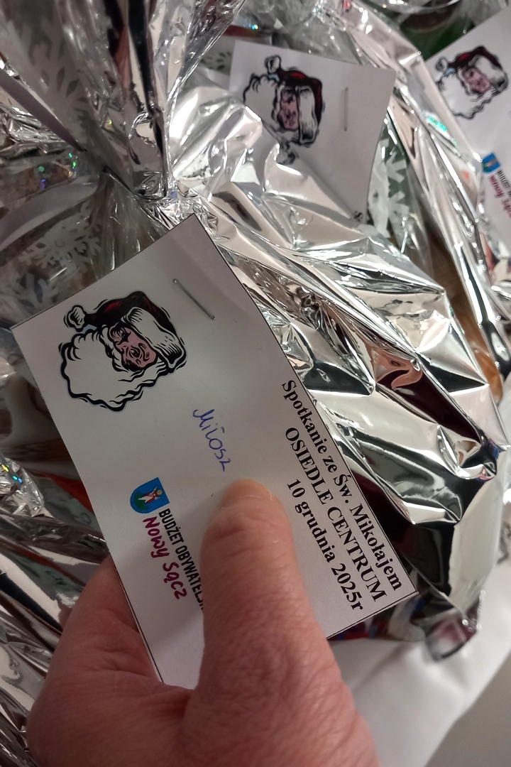 mikolajki12 Zbliżenie na karteczkę przypiętą do prezentu w srebrnym worku. Na kartce napis: spotkanie ze Świętym Mikołajem, Osiedle Centrum, 10 grudnia 2025 roku, ilustracja z głową Mikołaja, imię dziecka oraz herb miasta Nowego Sącza z napisem: Budżet Obywatelski Nowy Sącz.