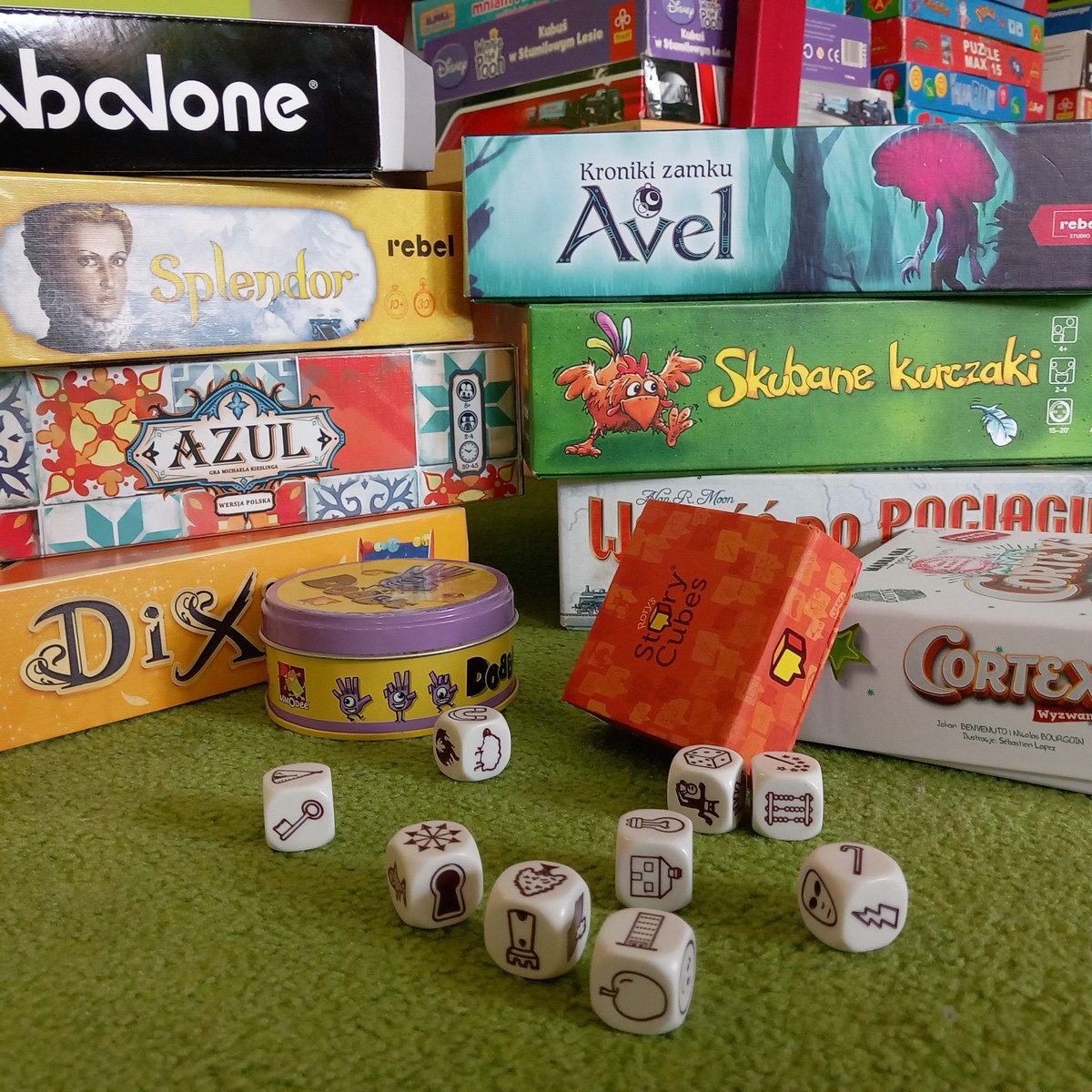 Zielony dywan. Na dywanie rozrzucone kostki do gry z symbolami. Za kostkami opakowania gier: dobble, story cubes, cortex. W tle stos różnych gier planszowych.