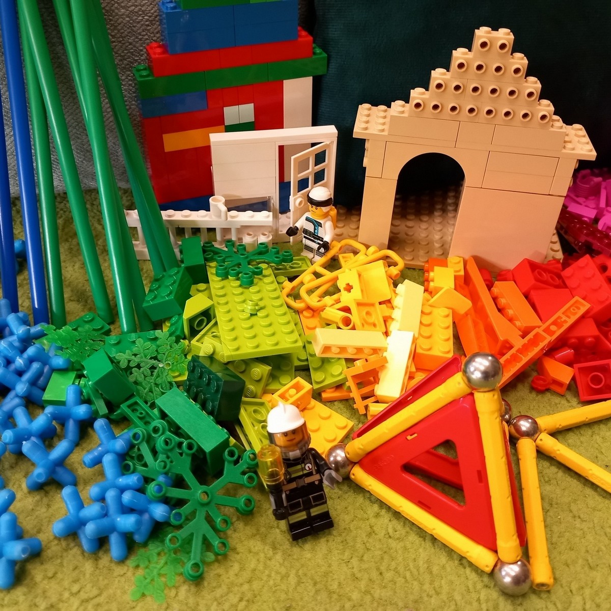 W pomieszczeniu na zielonym dywanie leżą zabawki: kolorowe klocki lego, rurki, łączenia do rurek, ludzik lego. Za leżącymi zabawkami przy ścianie stoją budowle domków i bramy z klocków lego.