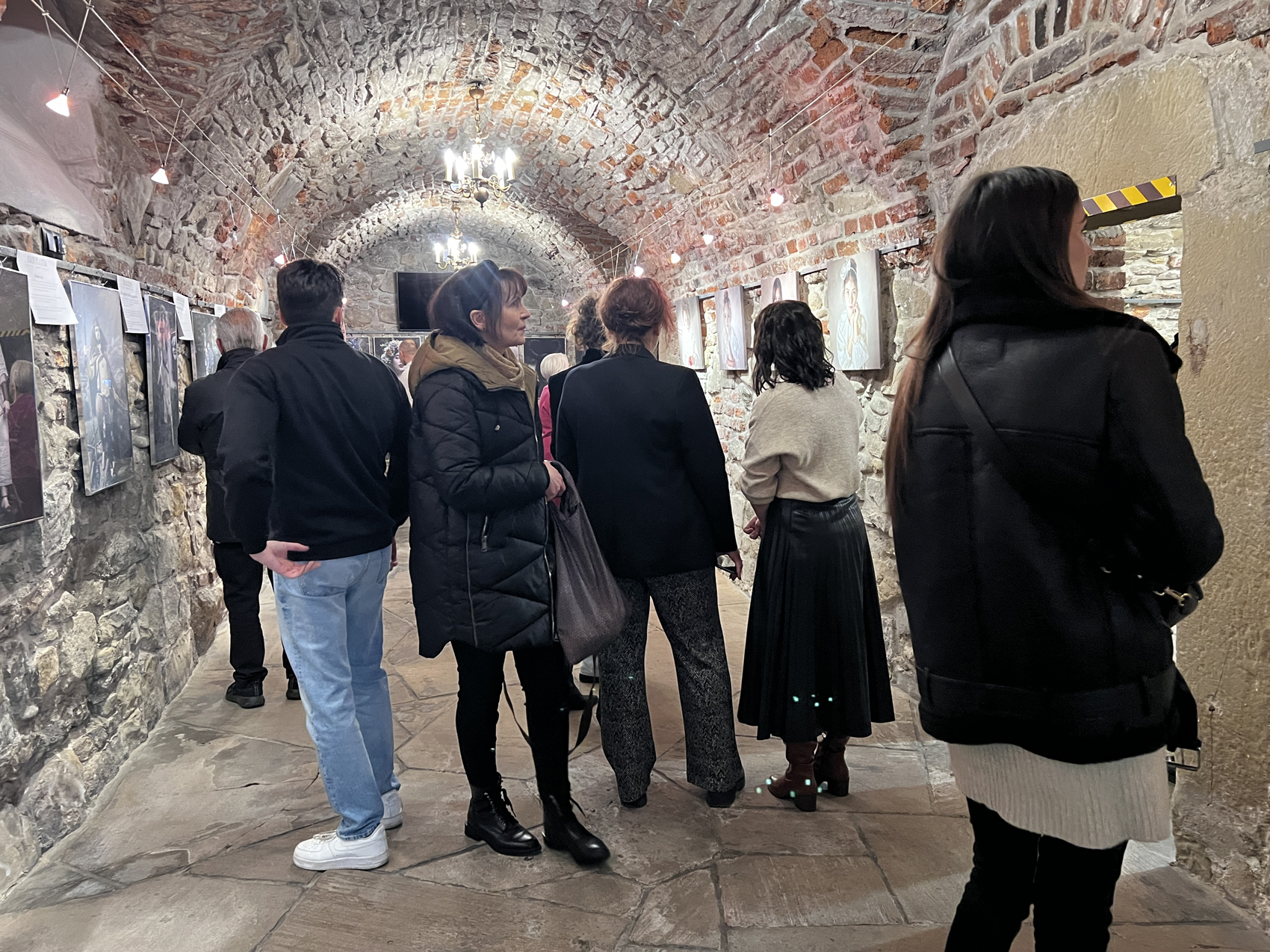 Finisaż absolwentów szkoły fotografii (1) Pomieszanie z zaokrąglonymi ceglanymi sufitami i kamiennymi ścianami i podłogami. Grupa ludzi stoi i patrzy na powieszone na ścianach zdjęcia. Ludzie ubrani są w kurtki i płaszcze.