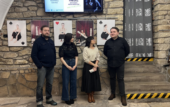 Cztery osoby - od lewej: mężczyzna w średnim wieku w ciemnych jeansach i granatowej kurtce; młoda dziewczyna w czarnych włosach do ramion, ma na sobie ciemne granatowe jeansy i czarną koronkową bluzkę. Patrzy w prawo; kobieta w czarnych włosach do ramion, ma okulary. Ubrana jest w beżowy sweterek z podwiniętymi rękawami i czarną plisowaną spódnicę do połowy łydki. Również patrzy w prawo. Obok niej stoi młody chłopak w czarnych jeansach i czarnej koszuli. Uśmiecha się. Wszyscy stoją na tle kamiennej ścianki z małym murkiem. Na ścianie wisi pięć zdjęć - trzy białe w stylu karcianym i dwa czarno-bordowe w tematyce szachów. Na każdym ze zdjęć znajduje się młoda kobieta. Wyżej widać fragment telewizora z grafiką i podpisem: finisaż wystawy absolwentów MASTERCLASS. Po prawej stronie widać kamienne schody i przejście zasłonięte szarą zasłoną.