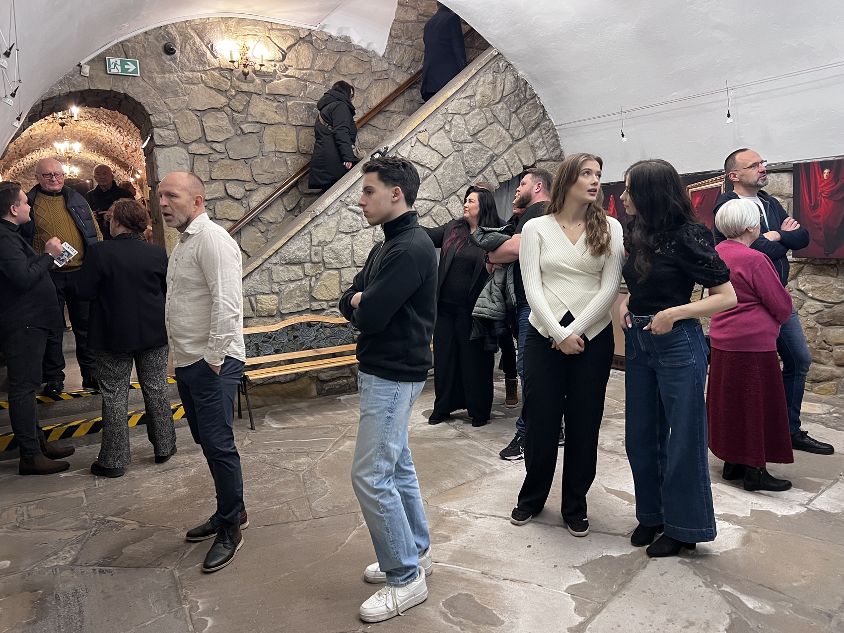 Finisaż absolwentów szkoły fotografii (3) Pomieszanie z zaokrąglonymi sufitami i kamiennymi ścianami i podłogami. Sufity są białe. Duża grupa ludzi stoi i patrzy w różne strony. Przy ścianie widać wysokie schody. Na ścianach wiszą czarno-czerwone obrazy.
