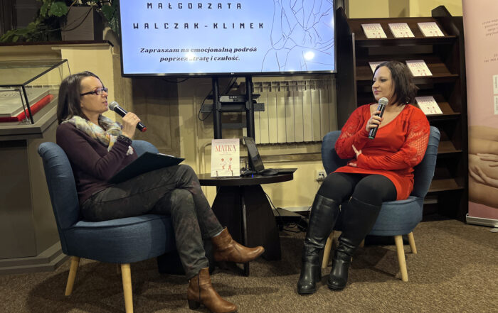 W pomieszczeniu na niskich niebieskich fotelach siedzą dwie kobiety. Każda w dłoni trzyma blisko ust mikrofon. Patrzą na siebie i rozmawiają ze sobą. Kobieta od lewej ubrana jest w brązową bluzkę z długim rękawem, kolorową apaszkę, ciemne spodnie i brązowe botki. Kobieta po prawej ubrana jest w czerwoną bluzkę z długim rękawem, czarne spodnie i czarne wysokie kozaki. Między siedzącymi kobietami stoi mały okrągły brązowy stolik a na nim otwarty laptop i książka z tytułem "Matka". W tle duży ekran z wyświetloną białą grafiką i czarnymi napisami: "Małgorzata Walczak-Klimek. Zapraszam na emocjonalną podróż poprzez stratę i czułość". Obok po prawej stronie wysoki brązowy regał z książkami.
