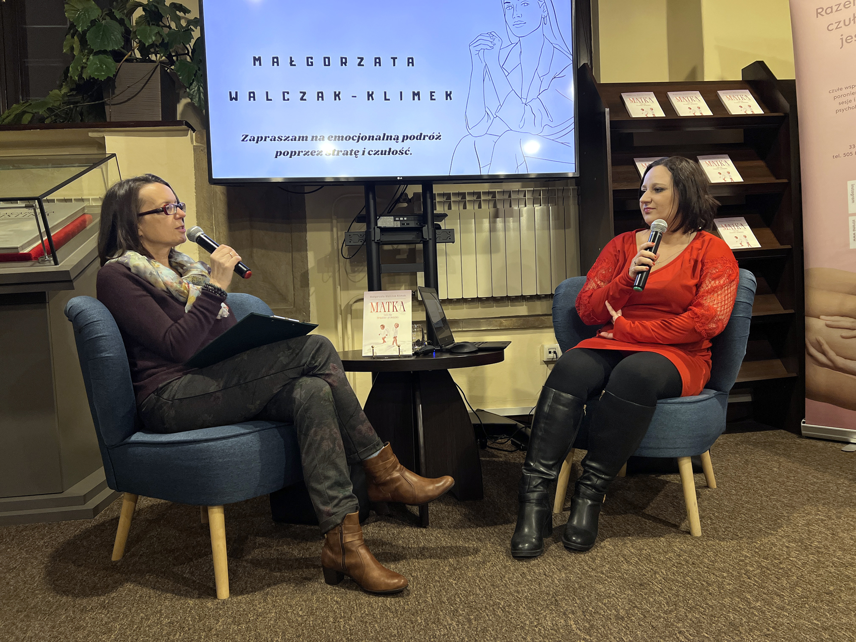W pomieszczeniu na niskich niebieskich fotelach siedzą dwie kobiety. Każda w dłoni trzyma blisko ust mikrofon. Patrzą na siebie i rozmawiają ze sobą. Kobieta od lewej ubrana jest w brązową bluzkę z długim rękawem, kolorową apaszkę, ciemne spodnie i brązowe botki. Kobieta po prawej ubrana jest w czerwoną bluzkę z długim rękawem, czarne spodnie i czarne wysokie kozaki. Między siedzącymi kobietami stoi mały okrągły brązowy stolik a na nim otwarty laptop i książka z tytułem "Matka". W tle duży ekran z wyświetloną białą grafiką i czarnymi napisami: "Małgorzata Walczak-Klimek. Zapraszam na emocjonalną podróż poprzez stratę i czułość". Obok po prawej stronie wysoki brązowy regał z książkami.