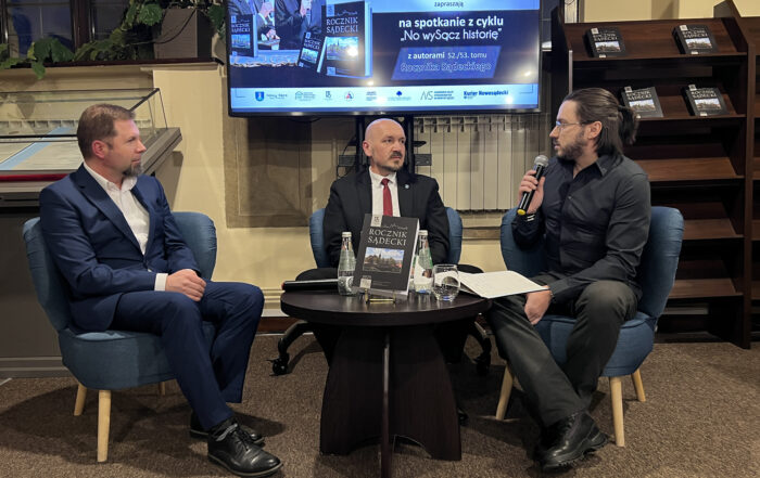 W pomieszczeniu czytelni na niskich fotelach siedzą trzej mężczyźni. Na środku między nimi stoi mały okrągły brązowy stolik. Na stoliku znajdują się: czarna okładka książki z tytułem "Rocznik Sądecki", dwie szklane butelki z wodą, szklanka, mikrofon, otwarty notes. Mężczyzna pierwszy od prawej ubrany jest w czarną koszulę i ciemne spodnie. W jednej ręce trzyma mikrofon blisko ust. Patrzy na mężczyznę siedzącego obok, ubranego w czarną marynarkę, czerwony krawat i białą koszulę. Mężczyzna siedzący od lewej ubrany jest w granatowy garnitur i białą koszulę. W tle stoi duży ekran z wyświetloną grafiką. Na grafice napisy: "spotkanie z cyklu No wySącz historię, z autorami 52./53. tomu Rocznika Sądeckiego". Po prawej stronie ekranu stoi wysoki brązowy regał a na nim leżą cztery książki.