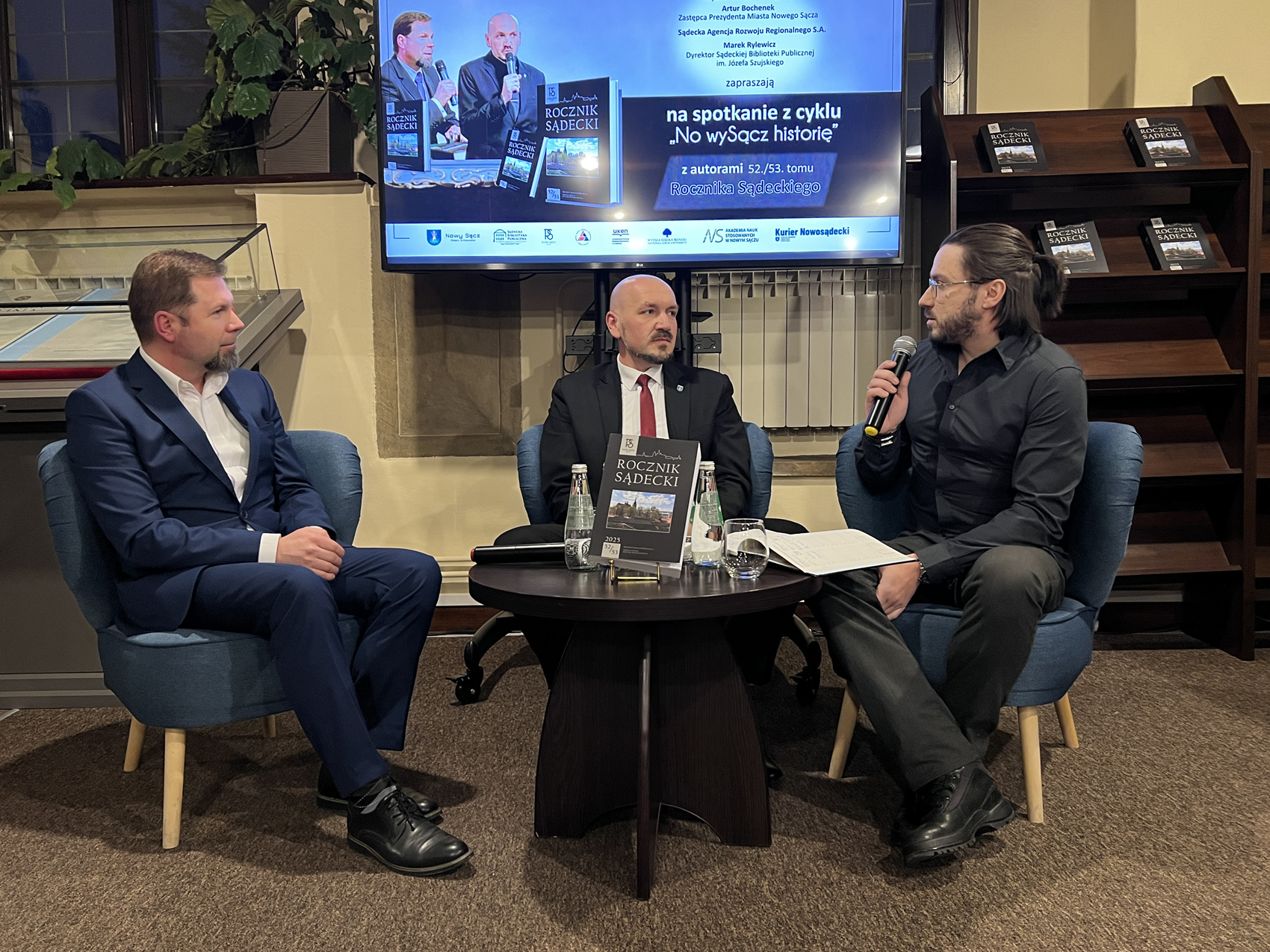 W pomieszczeniu czytelni na niskich fotelach siedzą trzej mężczyźni. Na środku między nimi stoi mały okrągły brązowy stolik. Na stoliku znajdują się: czarna okładka książki z tytułem "Rocznik Sądecki", dwie szklane butelki z wodą, szklanka, mikrofon, otwarty notes. Mężczyzna pierwszy od prawej ubrany jest w czarną koszulę i ciemne spodnie. W jednej ręce trzyma mikrofon blisko ust. Patrzy na mężczyznę siedzącego obok, ubranego w czarną marynarkę, czerwony krawat i białą koszulę. Mężczyzna siedzący od lewej ubrany jest w granatowy garnitur i białą koszulę. W tle stoi duży ekran z wyświetloną grafiką. Na grafice napisy: "spotkanie z cyklu No wySącz historię, z autorami 52./53. tomu Rocznika Sądeckiego". Po prawej stronie ekranu stoi wysoki brązowy regał a na nim leżą cztery książki.