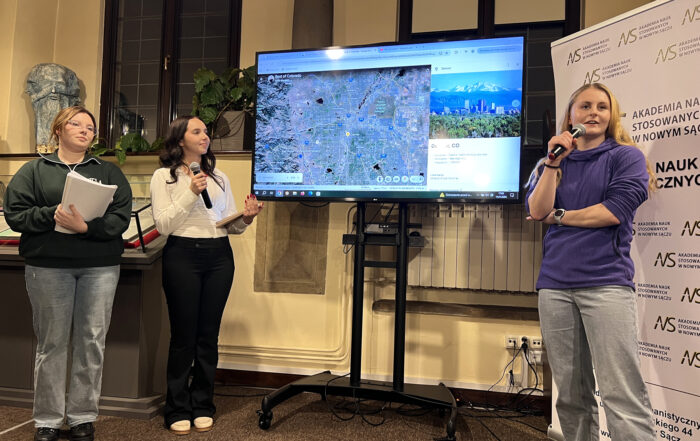 W pomieszczeniu czytelni stoją trzy młode dziewczyny. Dwie dziewczyny stoją po lewej stronie, jedna po prawej. Między nimi stoi duży ekran a nim wyświetlona mapa. Dziewczyna po prawej stronie ubrana jest w jeansy i fioletową bluzę. W prawej ręce trzyma mikrofon blisko ust, mówi do niego. Za nią stoi biały rollup z logotypem Akademii Nauk Stosowanych w Nowym Sączu. Po lewej stronie pierwsza z dziewczyn od lewej ubrana jest w ciemnozieloną bluzę i jeansy. W rękach trzyma plik kartek. Obok niej dziewczyna ubrana w czarne spodnie i białą bluzkę. W jednej ręce trzyma tablet, w drugiej mikrofon.