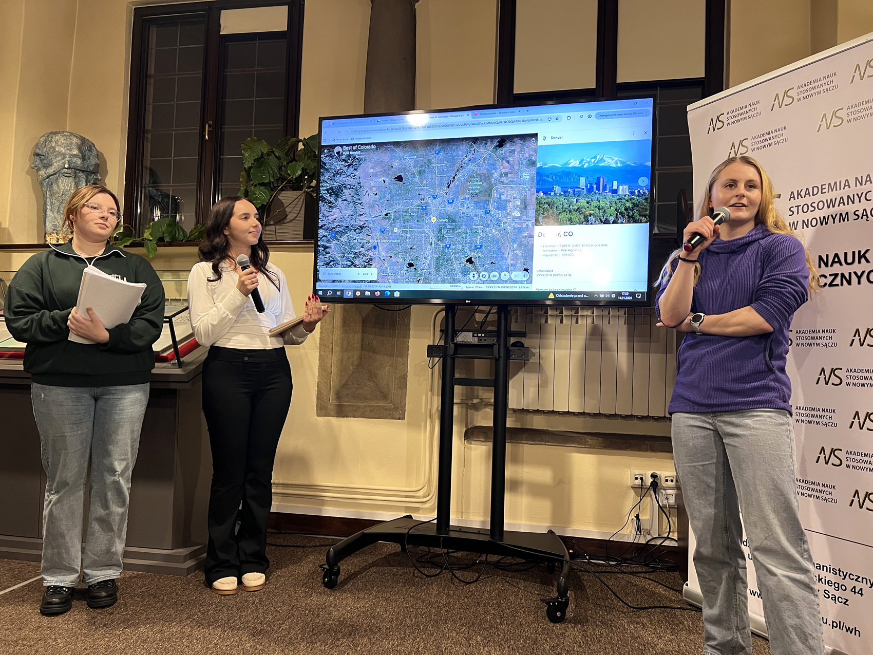 W pomieszczeniu czytelni stoją trzy młode dziewczyny. Dwie dziewczyny stoją po lewej stronie, jedna po prawej. Między nimi stoi duży ekran a nim wyświetlona mapa. Dziewczyna po prawej stronie ubrana jest w jeansy i fioletową bluzę. W prawej ręce trzyma mikrofon blisko ust, mówi do niego. Za nią stoi biały rollup z logotypem Akademii Nauk Stosowanych w Nowym Sączu. Po lewej stronie pierwsza z dziewczyn od lewej ubrana jest w ciemnozieloną bluzę i jeansy. W rękach trzyma plik kartek. Obok niej dziewczyna ubrana w czarne spodnie i białą bluzkę. W jednej ręce trzyma tablet, w drugiej mikrofon.