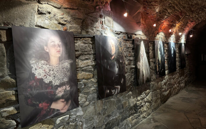 Długie pomieszczenie z kamienną ścianą, kamienną podłogą i półokrągłym sklepieniu z cegieł. Na ścianach wiszą obrazy. Nad każdym obrazem zapalona lampka.