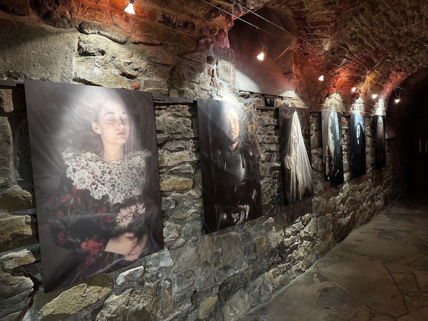 Długie pomieszczenie z kamienną ścianą, kamienną podłogą i półokrągłym sklepieniu z cegieł. Na ścianach wiszą obrazy. Nad każdym obrazem zapalona lampka.
