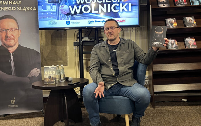 W pomieszczeniu na niskim fotelu siedzi mężczyzna ubrany w jeansy, czarną koszulkę i ciemnozieloną koszulę. Unosi lewą rękę, trzymając w niej książkę z czarną okładką i napisem "Uznanie". Po lewej stronie stoi niski okrągły brązowy stolik. Na stoliku znajdują się: dwie szklane butelki z wodą, mikrofon, telefon i książka. W tle od lewej: fragment rollupu z portretem mężczyzny, fragment ekranu z wyświetloną grafiką, fragment brązowego regału z książkami.