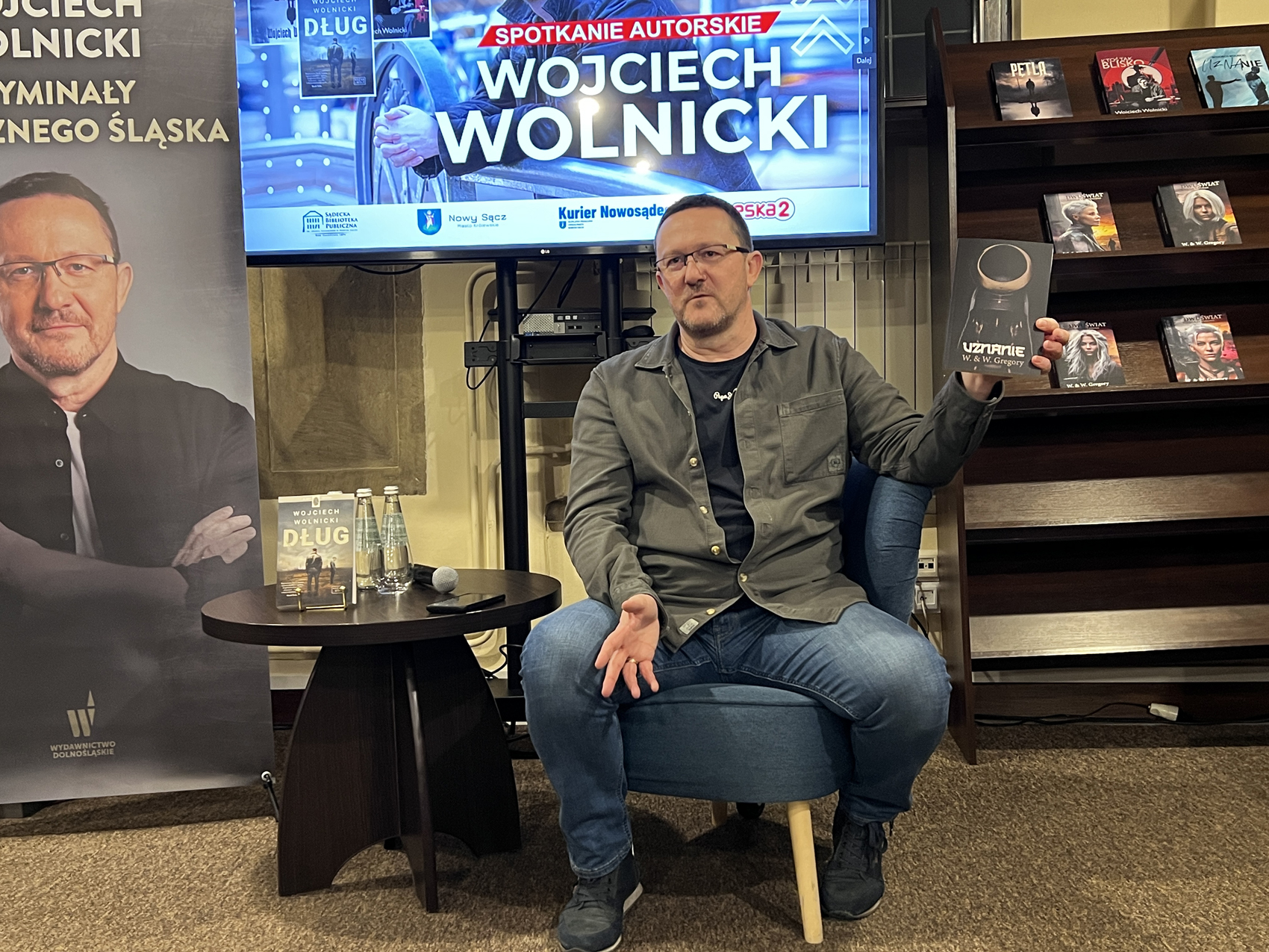 W pomieszczeniu na niskim fotelu siedzi mężczyzna ubrany w jeansy, czarną koszulkę i ciemnozieloną koszulę. Unosi lewą rękę, trzymając w niej książkę z czarną okładką i napisem "Uznanie". Po lewej stronie stoi niski okrągły brązowy stolik. Na stoliku znajdują się: dwie szklane butelki z wodą, mikrofon, telefon i książka. W tle od lewej: fragment rollupu z portretem mężczyzny, fragment ekranu z wyświetloną grafiką, fragment brązowego regału z książkami.