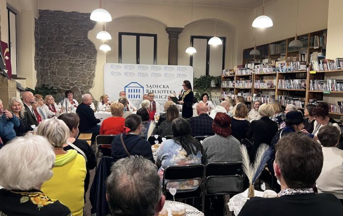 Duża sala w zabytkowym budynku z fragmentem kamiennej ściany, kamienną kolumną i dużymi oknami. Na sali jest tłum ludzi, którzy siedzą na krzesełkach przy małych okrągłych stolik lub większych na froncie. Pod oknami siedzi gruba starszych osób w białych koszulach i czerwonej przepasce na szyi. Za nimi stoi ścianka Sądeckiej Biblioteki Publicznej. W tłumie stoi jedna osoba, która trzyma w ręce mikrofon i podstawkę pod dokumenty. Po prawej stronie stoją regały pełne książek. Z sufity zwisa sześć zapalonych lamp.