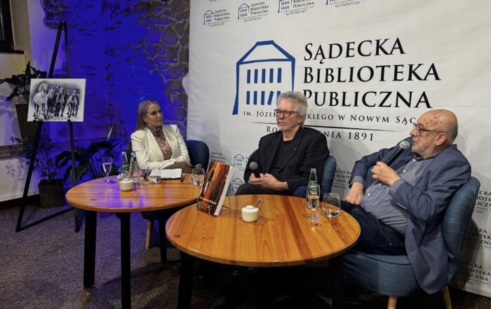 W pomieszczeniu na niskich fotelach siedzi jedna kobieta i dwóch starszych mężczyzn. Przed nimi stoją dwa okrągłe jasnobrązowe stoliki. Na stoliku pierwszym od lewej znajdują się: dwie szklane butelki z wodą, dwie szklanki, kieliszek szampana, biała cukiernica, podkładka z białą kartką. Na drugim stoliku znajdują się: szklanka, szklana butelka z wodą, kieliszek z szampanem, biała cukiernica i złota podkładka pod książkę a na niej książka. Kobieta siedzi po lewej stronie. Ubrana jest w białą marynarkę. Mężczyzna siedzący na środku ubrany jest w czarną marynarkę. W rękach opartych o kolana trzyma mikrofon. Mężczyzna od prawej ubrany jest w ciemne jeansy, niebieską koszulę i granatową marynarkę. W jednej ręce trzyma mikrofon blisko ust. W lewej części, obok kobiety, stoi czarna sztaluga a na niej czarno-biała grafika stada koni. Za sztalugą w rogu pomieszczenia niebiesko-fioletowe światło. Za siedzącymi ludźmi stoi biała wysoka ścianka reklamowa z logotypem Sądeckiej Biblioteki Publicznej.