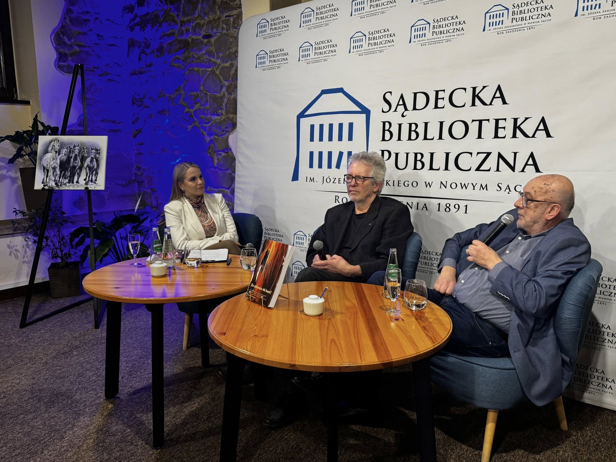 W pomieszczeniu na niskich fotelach siedzi jedna kobieta i dwóch starszych mężczyzn. Przed nimi stoją dwa okrągłe jasnobrązowe stoliki. Na stoliku pierwszym od lewej znajdują się: dwie szklane butelki z wodą, dwie szklanki, kieliszek szampana, biała cukiernica, podkładka z białą kartką. Na drugim stoliku znajdują się: szklanka, szklana butelka z wodą, kieliszek z szampanem, biała cukiernica i złota podkładka pod książkę a na niej książka. Kobieta siedzi po lewej stronie. Ubrana jest w białą marynarkę. Mężczyzna siedzący na środku ubrany jest w czarną marynarkę. W rękach opartych o kolana trzyma mikrofon. Mężczyzna od prawej ubrany jest w ciemne jeansy, niebieską koszulę i granatową marynarkę. W jednej ręce trzyma mikrofon blisko ust. W lewej części, obok kobiety, stoi czarna sztaluga a na niej czarno-biała grafika stada koni. Za sztalugą w rogu pomieszczenia niebiesko-fioletowe światło. Za siedzącymi ludźmi stoi biała wysoka ścianka reklamowa z logotypem Sądeckiej Biblioteki Publicznej.