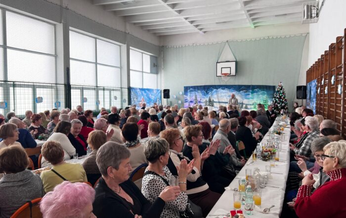 Sala gimnastyczna w szkole wypełniona ludźmi. Na środku ułożone są w rzędzie stoły z krzesłami. Ludzie siedzą przy stołach. Na stołach znajdują się: ciasta, soki, woda, szklanki. W tle na końcu sali zimowy krajobraz.