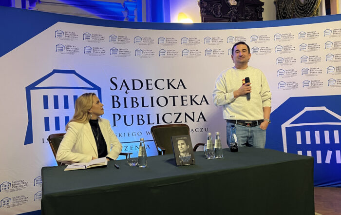 W pomieszczeniu stoi długi stół, nakryty zielonym obrusem. Na stole znajdują się: książka, trzy szklane butelki z wodą, dwie szklanki. Po lewej stronie na krześle przy stole siedzi młoda kobieta ubrana w czarny golf i białą marynarkę. Patrzy na stojącego obok niej mężczyznę. Po prawej stronie za stołem stoi mężczyzna ubrany w szarą bluzę i jeansy. W jednej ręce trzyma mikrofon. Za nimi w tle stoi biało-niebieska ścianka z logotypem Sądeckiej Biblioteki Publicznej.