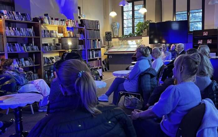 Sala czytelni wypełniona siedzącymi na krzesłach ludźmi, zwróconymi w lewą stronę. Przed nimi, po lewej stronie na dwóch niskich fotelach siedzą dwie kobiety. Za kobietami stoi długi regał z kolorowymi okładkami książek i audiobooków. Czytelnia podświetlona jest ciemnoniebieskim światłem.