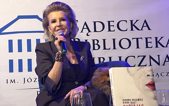 Na niskim fotelu siedzi kobieta ubrana w ciemnobrązowy garnitur. W prawej ręce trzyma mikrofon blisko ust. Obok niej siedzi mały kudłaty pies. Przed nią fragment okrągłego stolika. Na stoliku znajdują się dwie szklanki i książką z napisami: "Joanna Racewicz, Kiara Ulli, Sypiając z prezesem". W tle ścianka reklamowa Sądeckiej Biblioteki Publicznej.