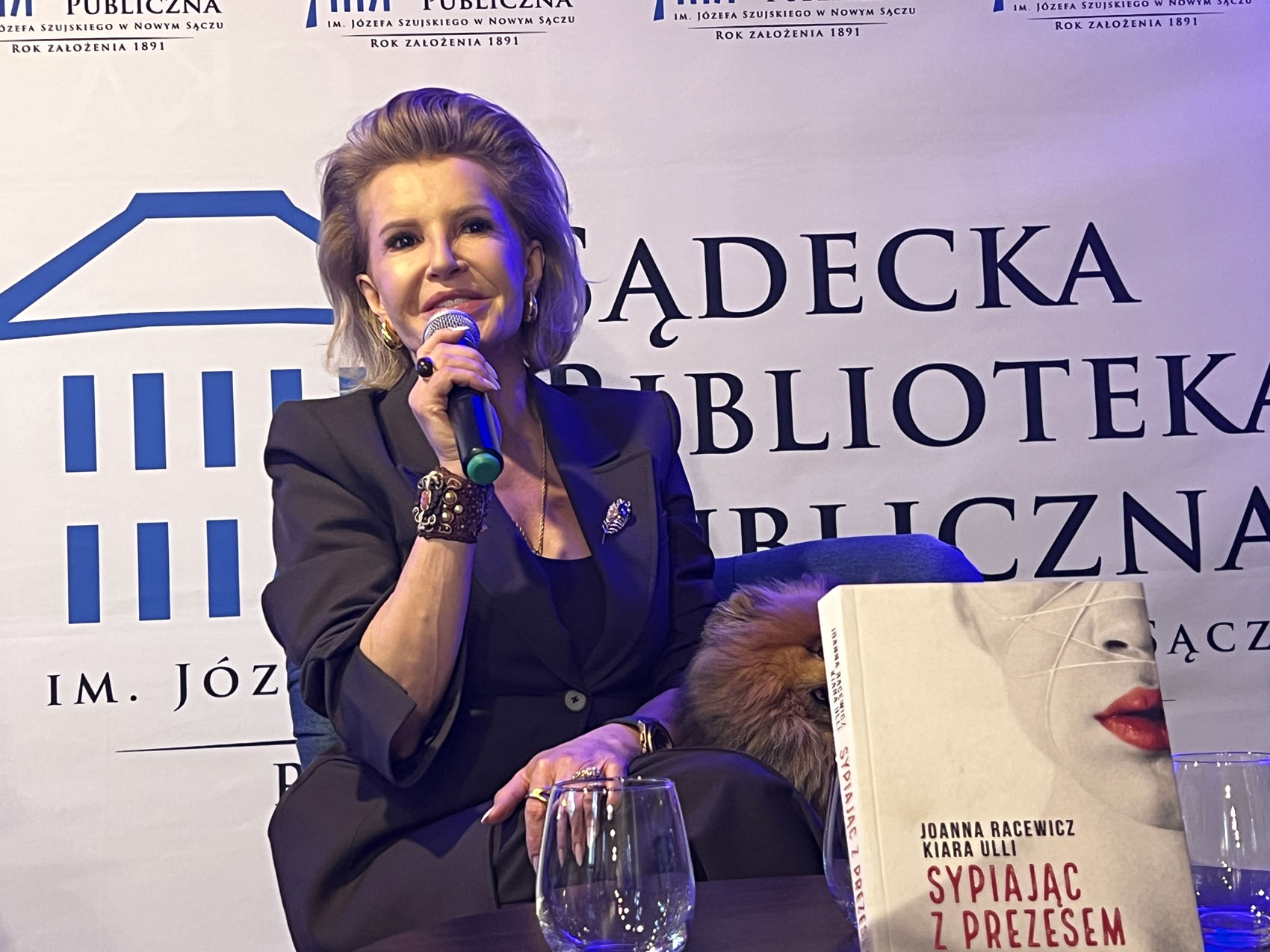 Na niskim fotelu siedzi kobieta ubrana w ciemnobrązowy garnitur. W prawej ręce trzyma mikrofon blisko ust. Obok niej siedzi mały kudłaty pies. Przed nią fragment okrągłego stolika. Na stoliku znajdują się dwie szklanki i książką z napisami: "Joanna Racewicz, Kiara Ulli, Sypiając z prezesem". W tle ścianka reklamowa Sądeckiej Biblioteki Publicznej.